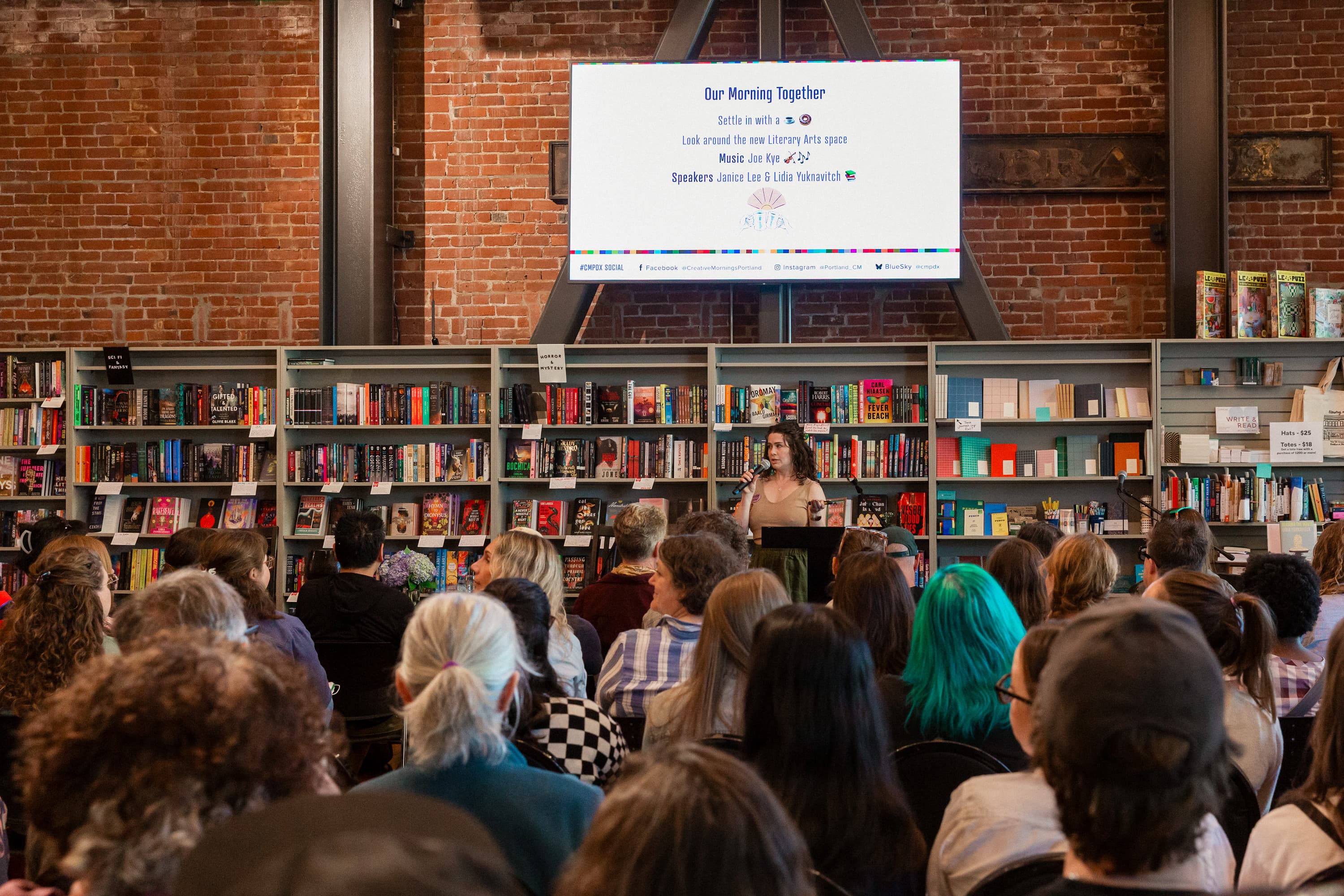 Kaitlin stands in front of a wall of bookshelves inside Literary Arts, speaking to a large seated crowd